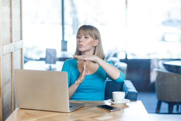 Portrait of hopeful young woman with blonde hair in blue shirt working on laptop, showing timeout gesture, asking about break, pleading. Indoor shot in cafe with big window on background.