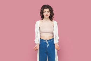 Portrait of unhappy sad upset woman with curly hair wearing casual style outfit standing looking away with puzzled expression, asking why. Indoor studio shot isolated on pink background.