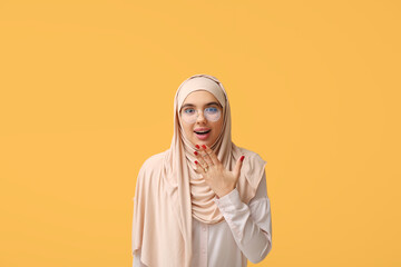 Shocked young Muslim woman in hijab and eyeglasses on yellow background