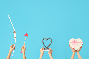 Female hands holding Cupid's bow, arrow, gift box and heart on blue background. Valentine's Day celebration