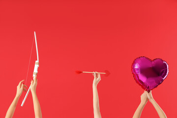 Female hands holding Cupid's bow, arrow and heart-shaped balloon on red background. Valentine's Day...
