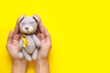 Female hands with toy bear and golden awareness ribbon on yellow background. International...