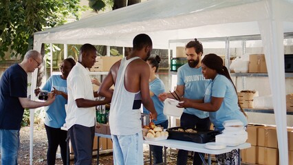 Male and female volunteers distribute prepared meals to homeless people and provide humanitarian aid. This scenario shows the significance of community solidarity and helping those in need. - obrazy, fototapety, plakaty