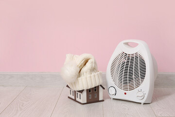 Decorative house with hat and electric fan heater near pink wall. Winter heating concept