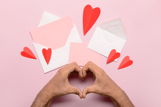 Male hands making heart shape with envelope, blank card and red paper hearts on pink background. Valentine's Day celebration