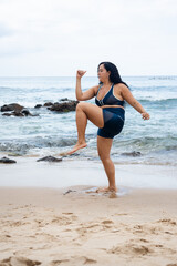 Fototapeta premium Fitness woman, standing, doing leg exercises on the beach sand against the sea and cloudy sky.