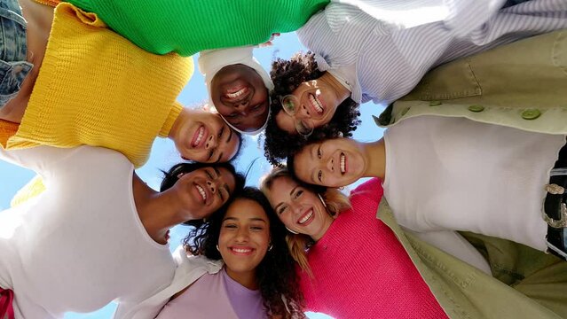Low angle view of diverse group of young women standing together in circle, hugging each other while smiling at camera. Female friendship and community, feminism and unity concept.