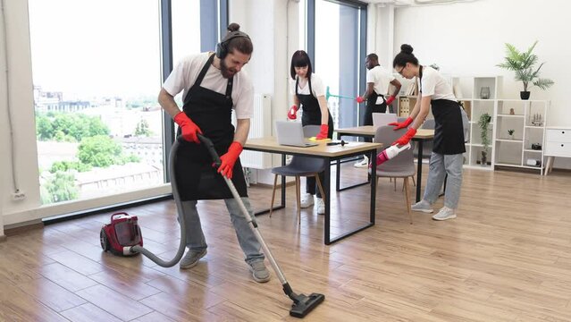 Professional cleaner Caucasian man having fun holding vacuum cleaner like guitar imitating playing at concert . Happy multinational team of people in black aprons cleaning modern office.