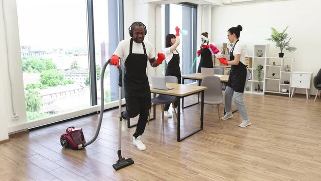 Adult bearded African man in headphones with modern vacuum cleaner and three multiracial cleaners in uniform listening music and dancing with equipment for cleaning on background of modern office.