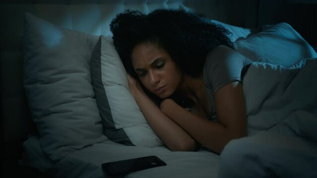 Beautiful African american girl sleeping in bed at home waking up turn off triggered alarm clock mobile phone message annoyed angry ethnic woman awake at night disturbed with incoming smartphone call