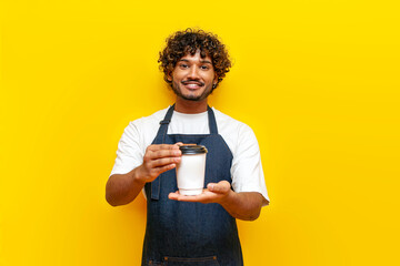young indian male barista in an apron holds and gives a paper glass of coffee on a yellow isolated...