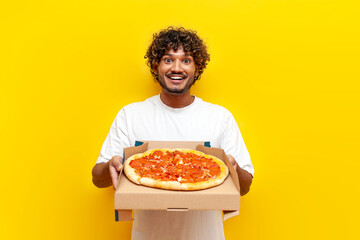 young hungry indian guy holding a box of delicious pizza and giving on a yellow isolated...