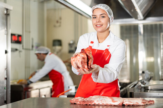 Smiling young girl in white uniform and red oilcloth apron working in meat processing room of butchery, standing near cutting table with fresh raw beef heart in hands..