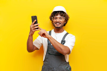 young indian male builder in a hard hat and overalls uses a smartphone and selects online on a...