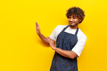 young indian male barista in an apron refuses and shows disgust on a yellow isolated background,...