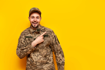 a young Ukrainian male soldier in a camouflage pixel uniform points his hand to the side on a yellow isolated background, the guy is a military cadet of the Ukrainian army advertising copy space - obrazy, fototapety, plakaty
