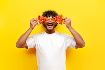 young funny indian guy closes his eyes with pieces of delicious pizza and smiles on yellow isolated background, man holds two pieces of pizza and advertises fast food - obrazy, fototapety, plakaty