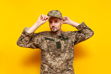 young Ukrainian male soldier in a pixel camouflage uniform puts on a cap on a yellow isolated...