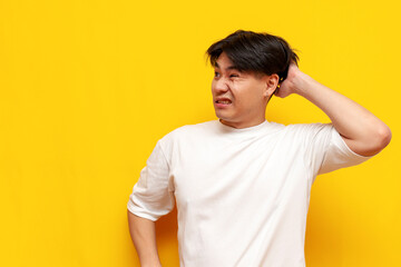 puzzled asian young man in white t-shirt thinks and remembers over yellow isolated background, confused korean guy student forgot and scratches head looking away