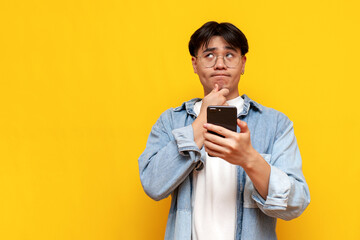 pensive asian young male student wearing glasses and denim shirt using smartphone and thinking on yellow isolated background, korean guy with phone planning and imagining looking away