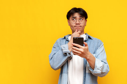 pensive asian young male student wearing glasses and denim shirt using smartphone and thinking on yellow isolated background, korean guy with phone planning and imagining looking away