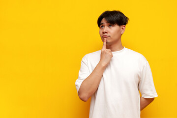 pensive asian young man in white shirt thinking and planning over yellow isolated background,...