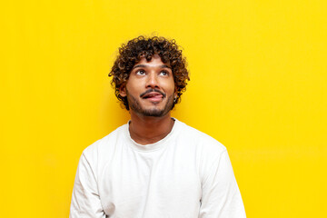 young curly indian man licking his lips and imagining on a yellow isolated background, a curious guy is dreaming and thinking looking up and showing his tongue - obrazy, fototapety, plakaty
