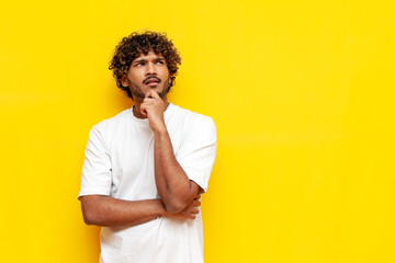 young pensive indian man in white t-shirt planning and thinking over yellow isolated background, curly puzzled guy imagining and dreaming. High quality photo