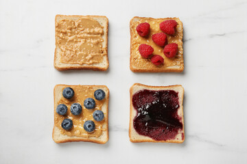 Delicious toasts with peanut butter, jam, raspberries and blueberries on white marble table, flat lay