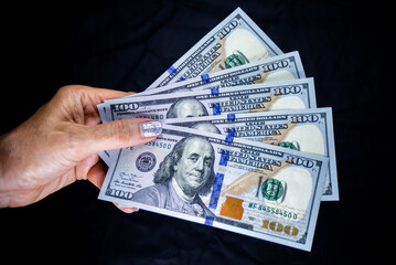 a woman's hand holding a fan of five 100 American dollar notes on a dark background	