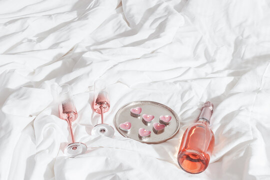Top view shiny champagne glasses, bottle of sparkling rose wine, pink candle hearts on white bed cloth. Minimal lifestyle aesthetic photo, romance mood, Valentine's Day, romantic love concept