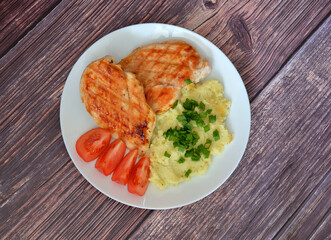 Two pieces of grilled chicken fillet with mashed potatoes and tomato slices on a wooden table.
