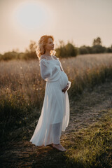 Fototapeta na wymiar A young expectant mother in the third trimester of pregnancy in a white dress hugs her stomach against the backdrop of a natural landscape. The concept of future motherhood.