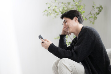 Profile of a man in long sleeves relaxing in a clean white living room looking at his phone.　Look...