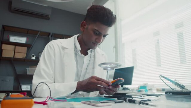 Medium shot of Indian male engineer in white coat using magnifying glass when repairing mobile phone at service centerMedium shot of Indian male engineer in white coat using magnifying glass when repa
