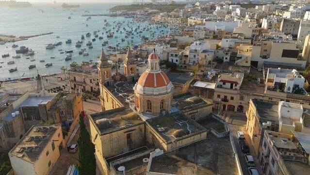 Aerial view of Marsaxlokk with lot of fishing boats in port and church at sunrise. Famous fishing village in Malta - Marsaxlokk. Morning UHD 4K shot