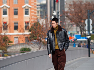 Obraz premium Portrait of handsome Chinese young man with black leather jacket posing with modern city street background in sunny winter day, male fashion, cool Asian young man.