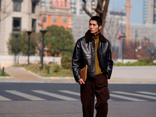 Portrait of handsome Chinese young man with black leather jacket posing with modern city street background in sunny winter day, male fashion, cool Asian young man.