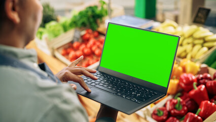 Anonymous African Farmer Working on a Computer with Green Screen Mock Up Display. Over the Shoulder Footage of a Street Vendor Using Laptop, Communicating with Sustainable Fruit and Vegetable Supplier - Powered by Adobe