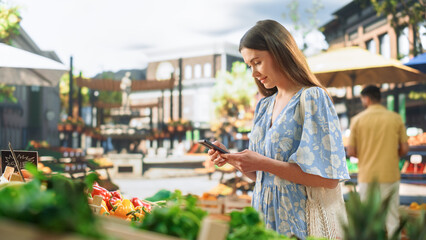 Customer in a Light Blue Dress Shopping for Fresh Seasonal Fruits and Vegetables, Using Smartphone to Browse Internet and Social Networks. Beautiful Woman Exploring a Farmers Market on Warm Sunny Day