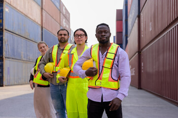 Team of multicultural engineers and workers in yellow vest standing together in shipping cargo container yard. International import and export logistic and transportation business concept. 
