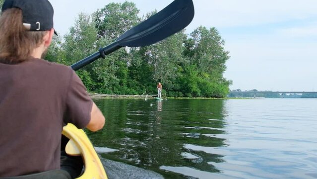 Man explores water by kayak during summer vacation. Sportsman swims to woman standing on sup board on lake among greenery