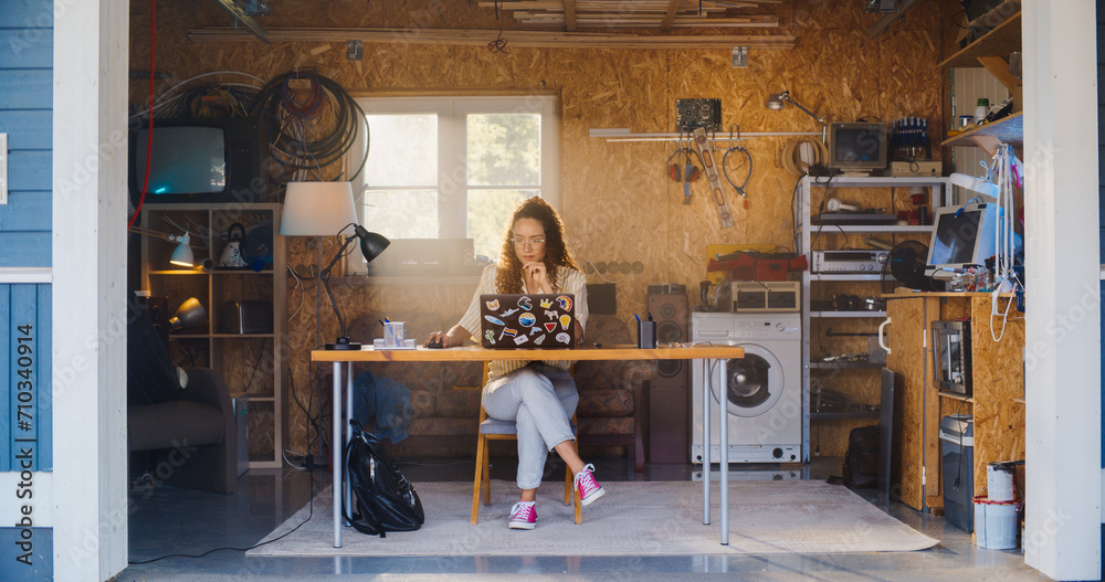 Poster hispanic female software developer coding on laptop computer in garage at home. focused young woman 