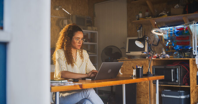 Hispanic Female Programmer Writing Code On Laptop Computer In Garage At Home. Woman Working On Innovative Software As A Service Startup For Business. Digital Entrepreneur Starting Company Concept.