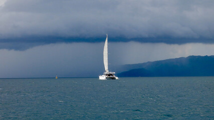 A catamaran with a raised sail sails across the blue sea towards the rain. A sailing catamaran moves across the sea among tropical islands towards a storm on a cloudy day.