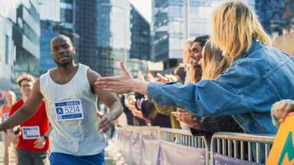 Marathon Audience Clapping and Cheering Their Loved Ones Participating in the Race: Black Athletic Male Marathon Runner Giving a High Five to Female Friend Supporting Him in the Audience While Running