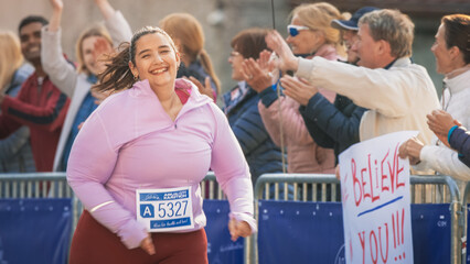 Portrait of a Smiling Plus Size Female Runner Crossing the Finish Line and Demonstrating her Willpower. Friendly City Marathon Audience Being Supportive - obrazy, fototapety, plakaty
