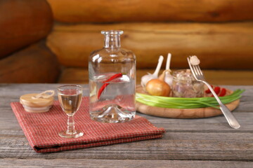 Obraz na płótnie Canvas On a plank wooden table against a log wall there is a glass of vodka in front of an open bottle next to a snack of pickled mushrooms and vegetables. 