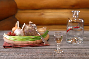 On a plank wooden table against a log wall there is a glass of vodka in front of an open bottle next to a snack of pickled mushrooms and vegetables. 