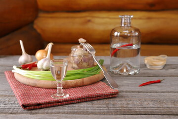 On a plank wooden table against a log wall there is a glass of vodka in front of an open bottle next to a snack of pickled mushrooms and vegetables. 